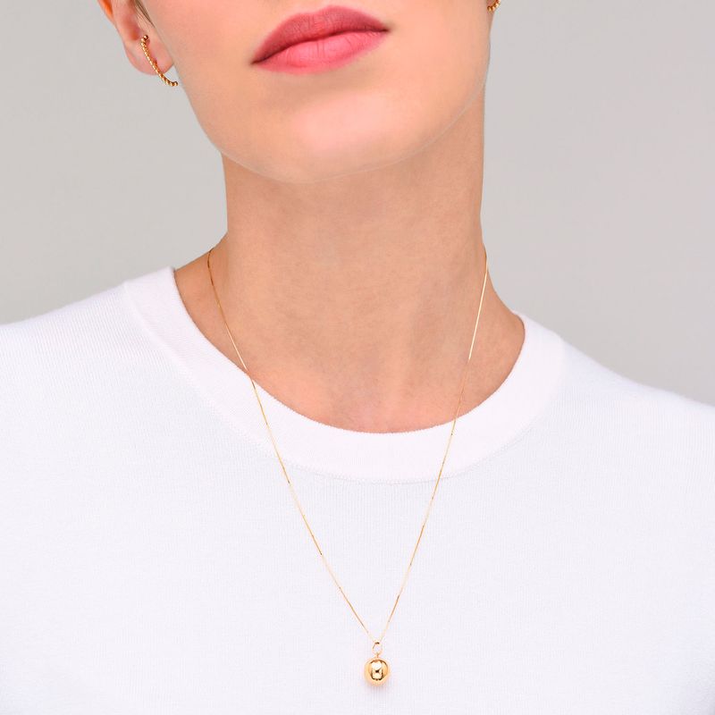 Colar dourado com pingente de esfera, usado por uma pessoa com camiseta branca.