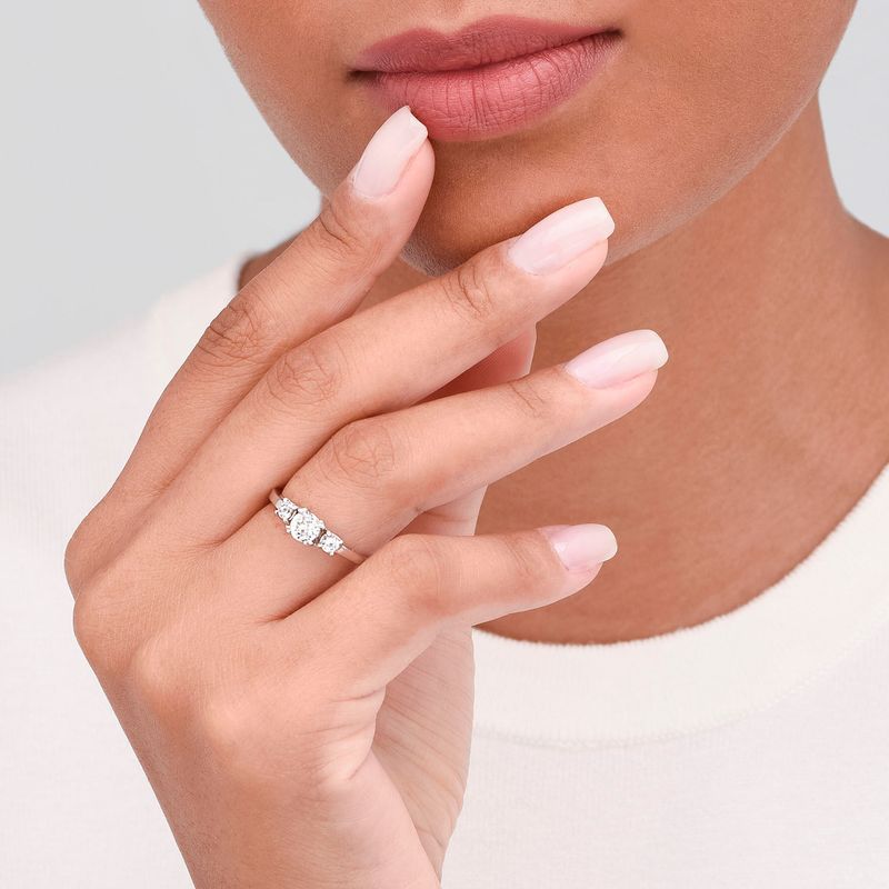 Mão de mulher usando anel de diamantes na mão esquerda, com lábios fechados e dedos próximos ao rosto.