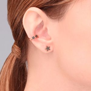 Conjunto de brincos com estrelas, um com pérola e spray de brilho preto, em orelha de pessoa com cabelo castanho.
