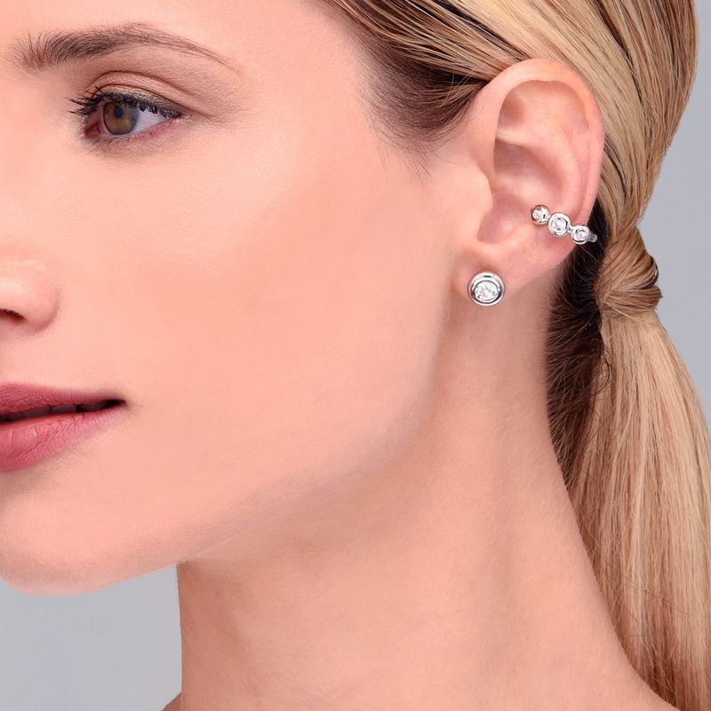 Close da orelha de uma mulher usando vários brincos de prata com pedras preciosas transparentes, incluindo um brinco de pino e um bracelete com três pedras.