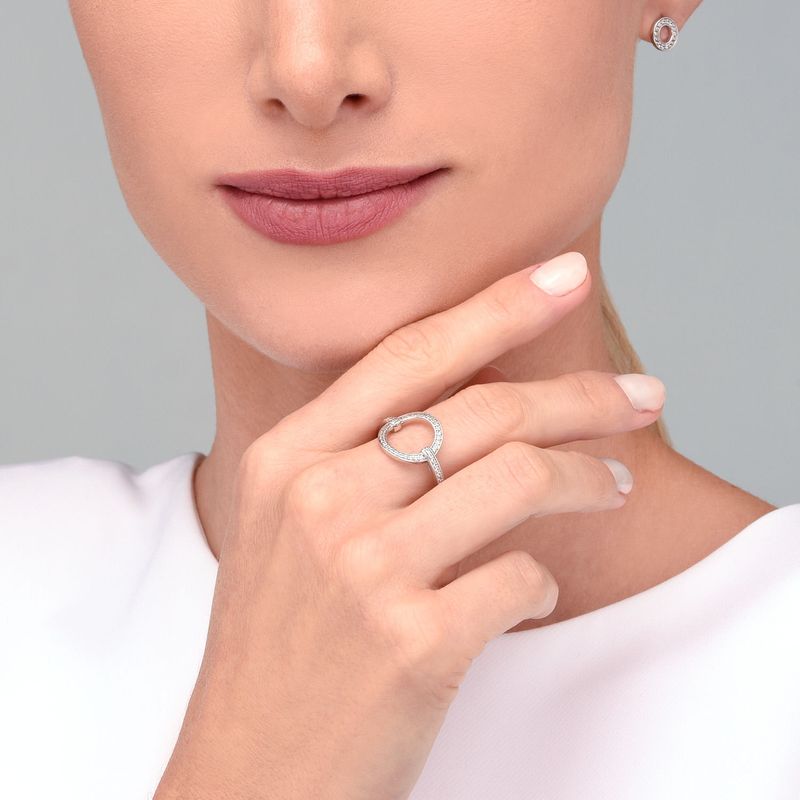 Mulher usando um anel de prata com design circular e detalhes de pedras, segurando o queixo com a mão direita.