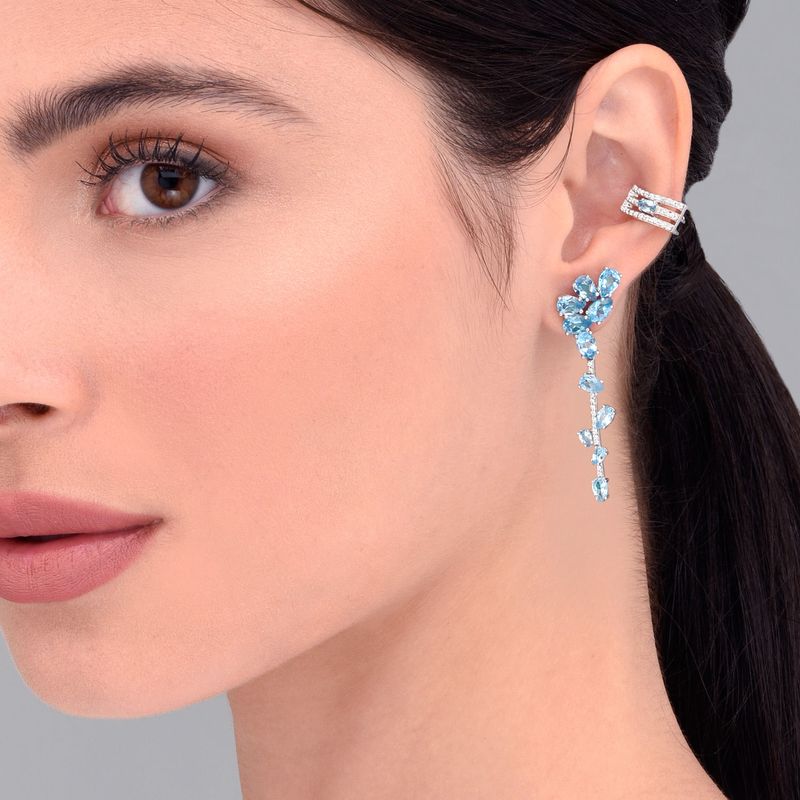 Close do rosto de uma mulher, mostrando seus olhos castanhos, lábios rosados ??e usando vários brincos, incluindo um longo brinco de pedra preciosa azul.