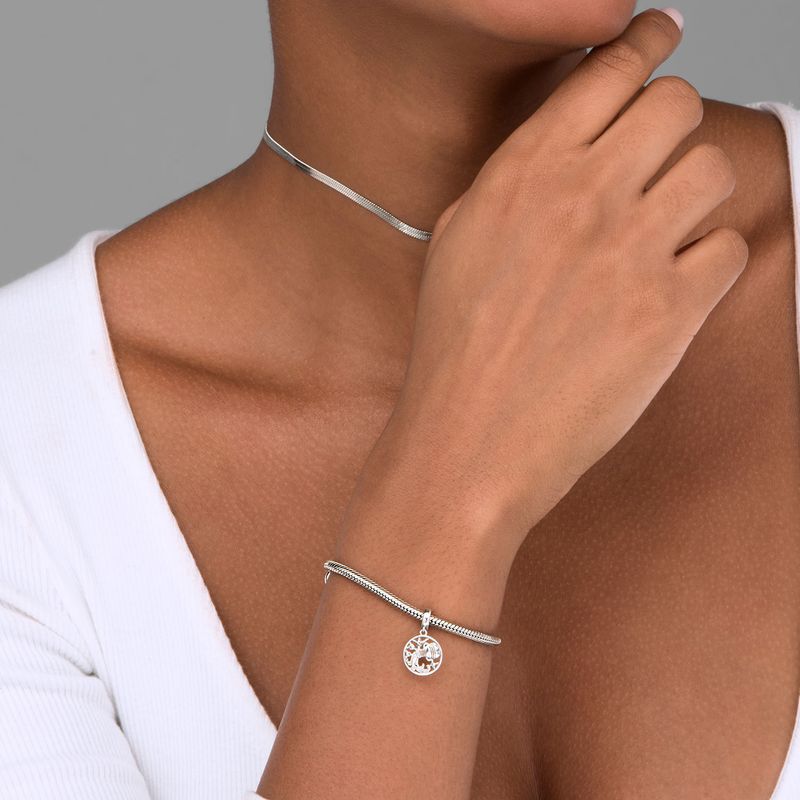 Mão e braço de uma mulher usando uma pulseira com pingente e um colar fininho, ambos de prateado, com pele morena e vestindo roupa branca.