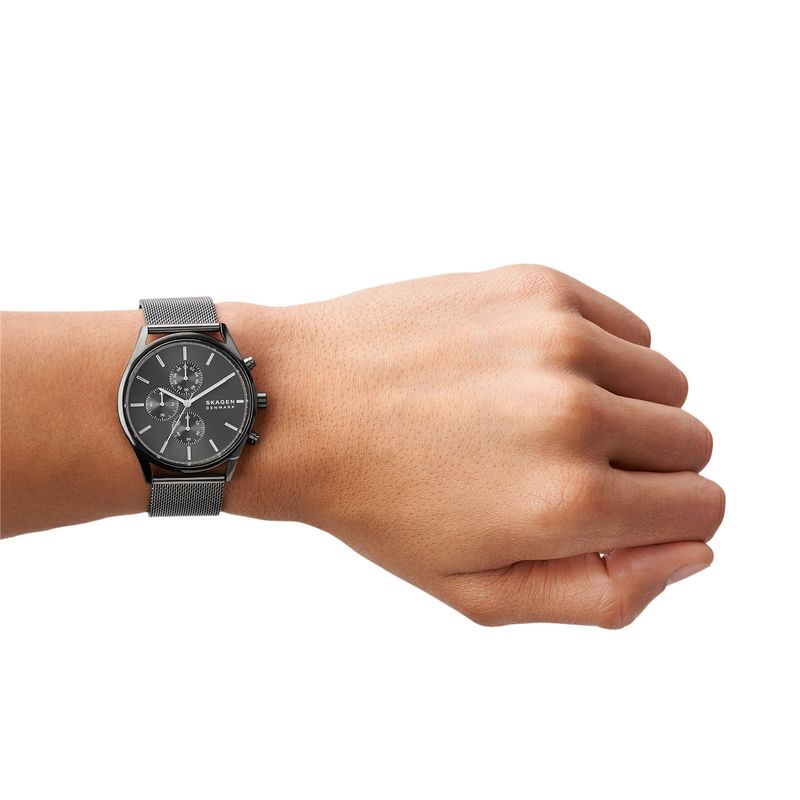 Relógio preto com pulseira de malha metálica no pulso de uma pessoa, com fundo branco.