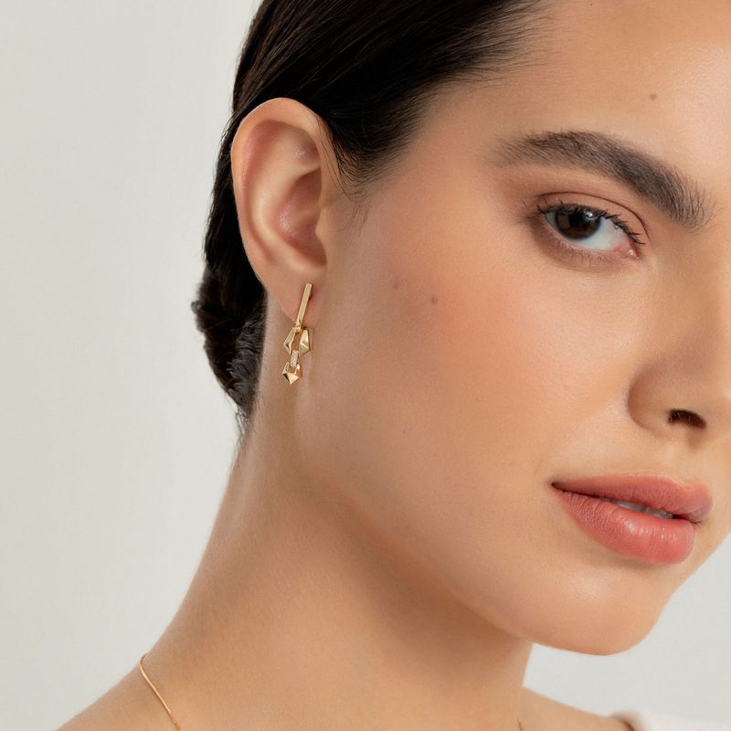 Close de uma mulher usando brincos de ouro com formas geométricas e um colar combinando contra um fundo claro.