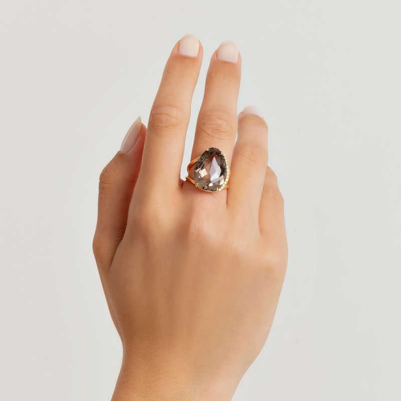Dedo com anel grande com pedra brilhante em formato de gota, em fundo branco.