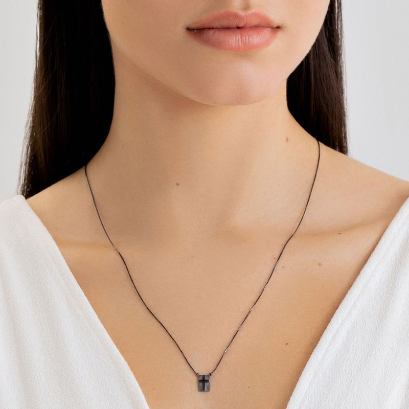 Colar fino de corrente preta com pingente quadrado preto, usado por uma mulher com blusa branca.
