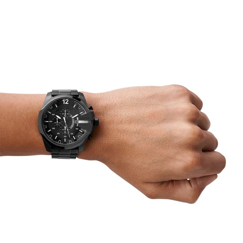 Relógio preto com pulseira de metal no pulso de uma pessoa, exibindo um mostrador escuro com detalhes em branco.
