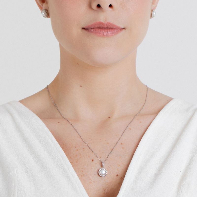 Colar delicado com pingente de pérola envolto por pequenos diamantes, usado por mulher com blusa branca.
