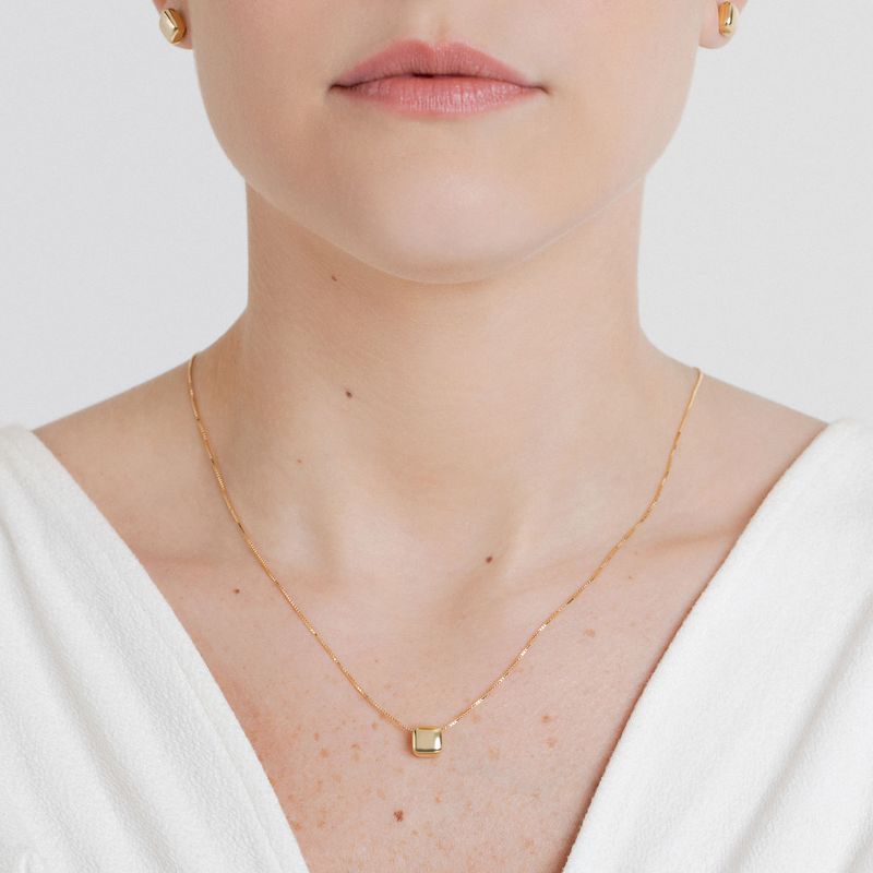 Gargantilha de corrente dourada com pingente quadrado dourado, sendo usada por uma mulher com pele clara e blusa branca.