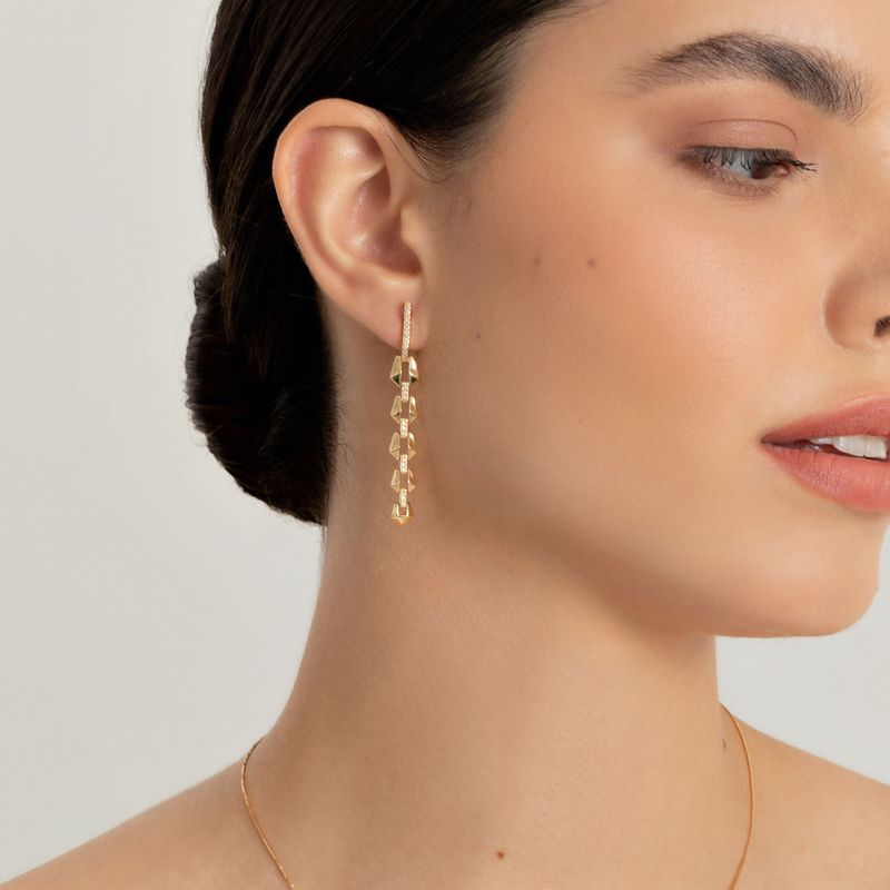 Close de uma mulher usando um brinco dourado pendurado com várias formas geométricas, junto com um delicado colar de ouro.