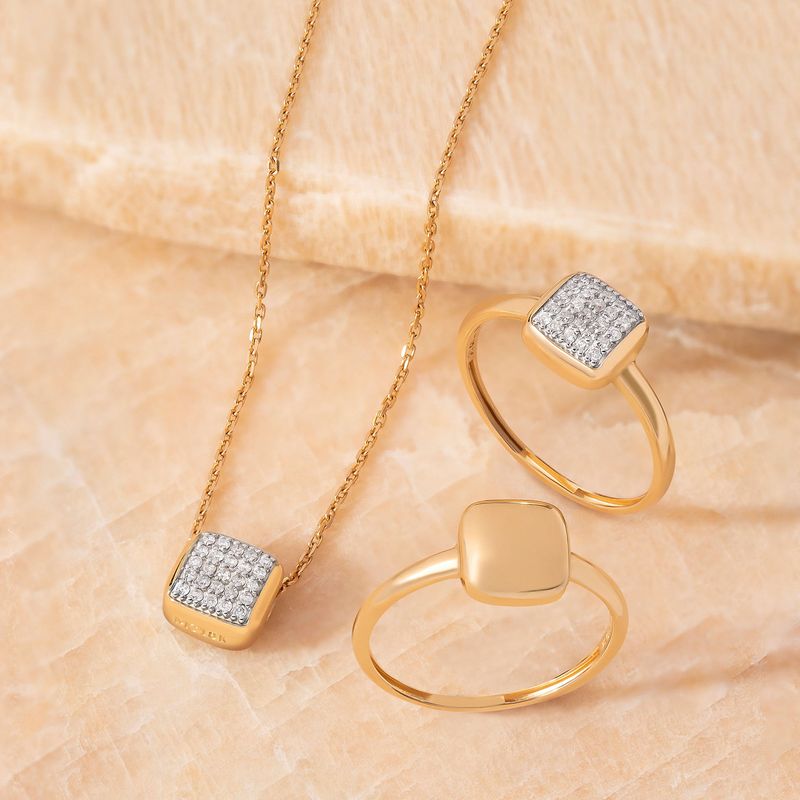Conjunto de joias douradas com pingente, anéis com pedras brilhantes e uma corrente, exibido sobre uma superfície de pedra beige.
