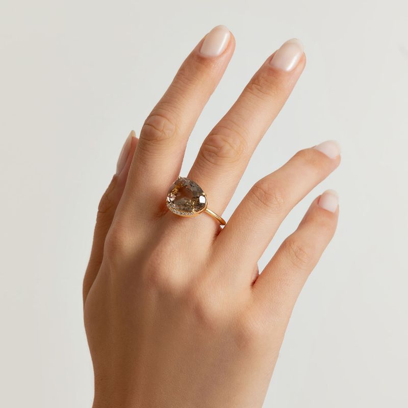 Anel com pedra grande em formato de coração, na cor marrom, no dedo de uma mão com unhas bem cuidadas.