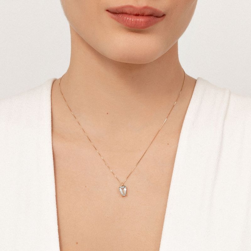 Colar dourado com pingente de pedra cristalina em formato retangular, usado por uma mulher com blusa branca.