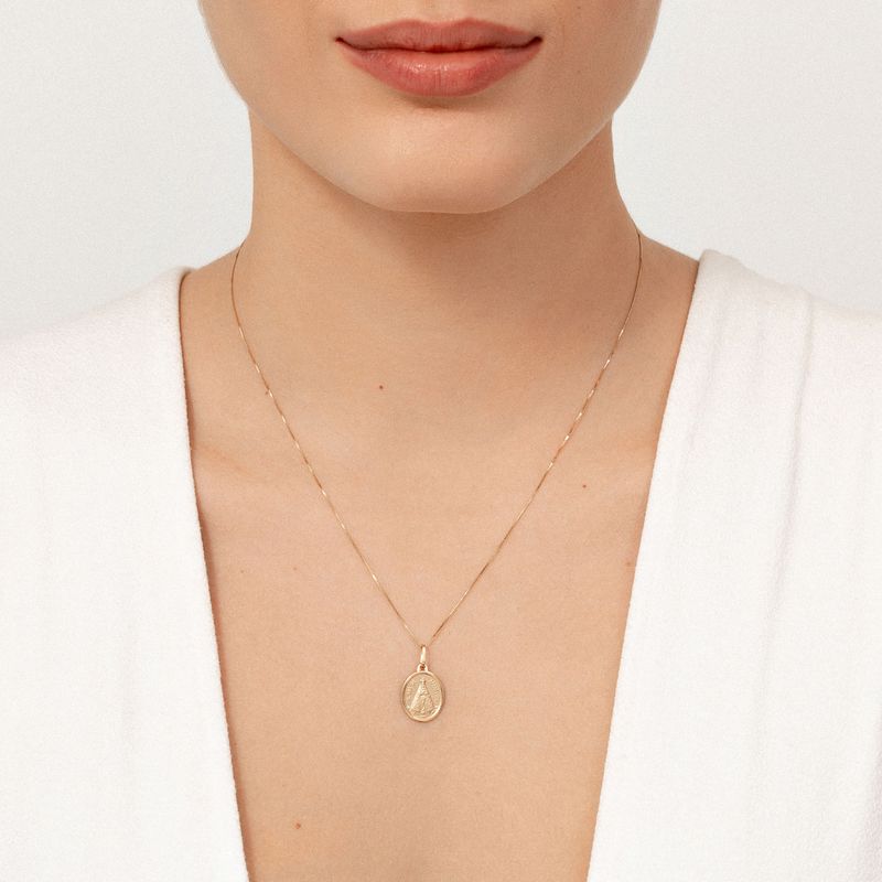 Colar dourado com pingente de medalha, usado por uma pessoa com blusa branca.