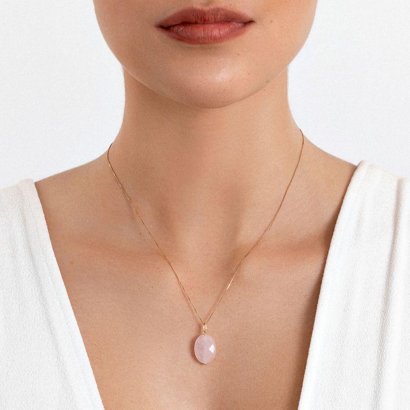 Colar delicado com pingente de pedra rosa oval, usado por uma pessoa com pele clara e blusa branca.