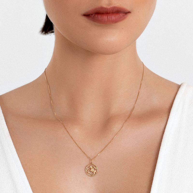 Colar dourado com pingente circular e desenho estilizado, usado por uma mulher com camisa branca e pele clara.
