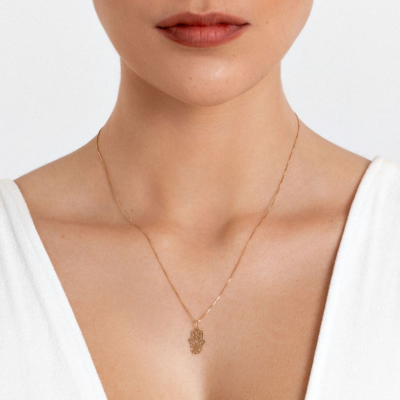Colar dourado com pingente de mão na frente, usado por uma pessoa com pele clara e vestindo blusa branca.