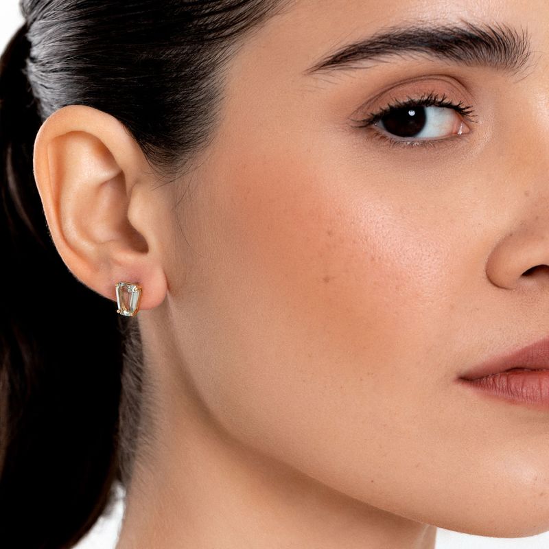 Close do rosto de uma mulher com foco em sua orelha com um brinco de ouro com uma pedra preciosa retangular transparente.