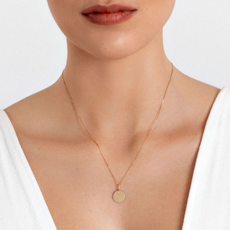 Colar delicado de corrente fina dourada com pingente circular liso, usado por uma mulher com roupa branca e pele clara.