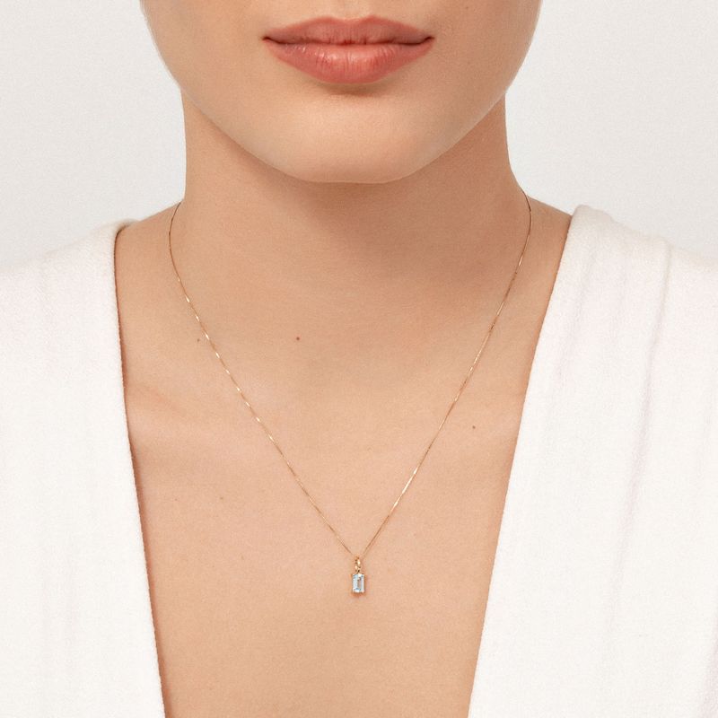 Colar dourado delicado com pingente de pedra azul e base de cadeia fina, usada no pescoço de uma mulher com roupa branca e pele clara.
