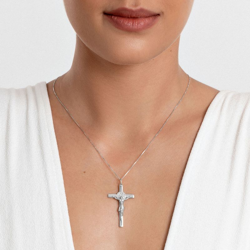 Colar com pingente de crucifixo de prata e detalhes em pedras transparente, usado por uma mulher de pele clara e roupas brancas.