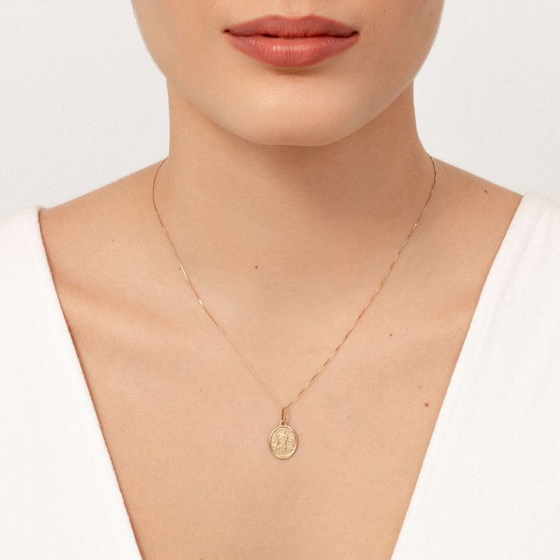 Colar delicado de corrente fina com medalha dourada oval com figura central, usado no pescoço de uma mulher.