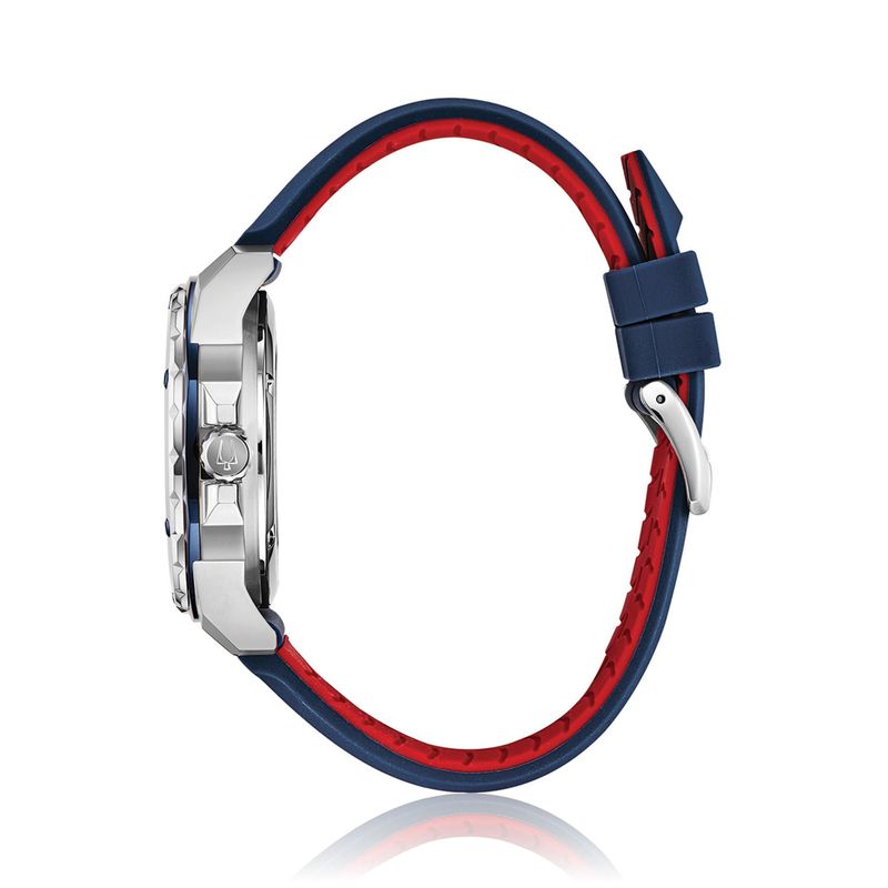 Relógio com pulseira azul e vermelha, caixa prateada, vista lateral mostrando o lado esquerdo, com botão de ajuste na lateral.