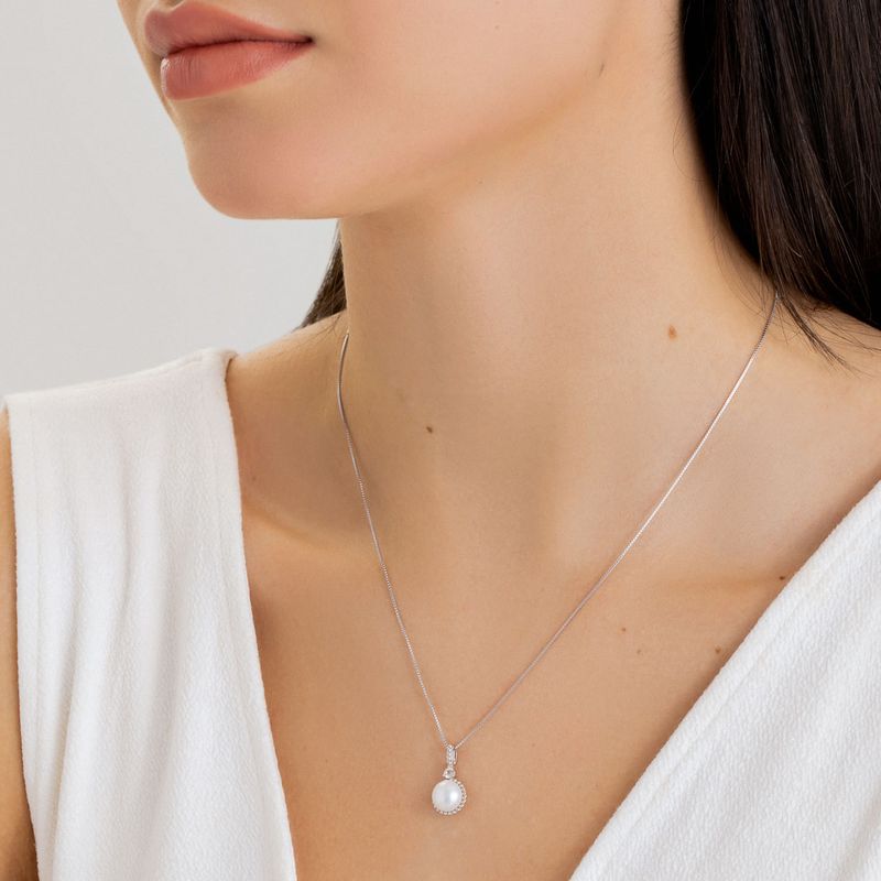 Colar delicado de prata com pingente de pérola e pequenos detalhes brilhantes, usado por uma mulher com pele clara e cabelo castanho escuro.
