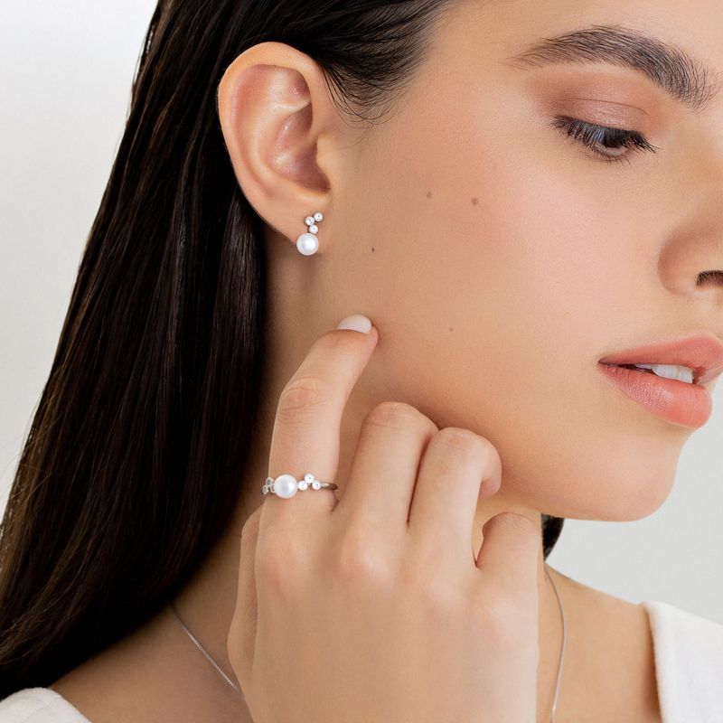 Mulher com brincos, anel e colar de pérolas elegantes, com pele clara e cabelo castanho escuro, posando com delicadeza na face.