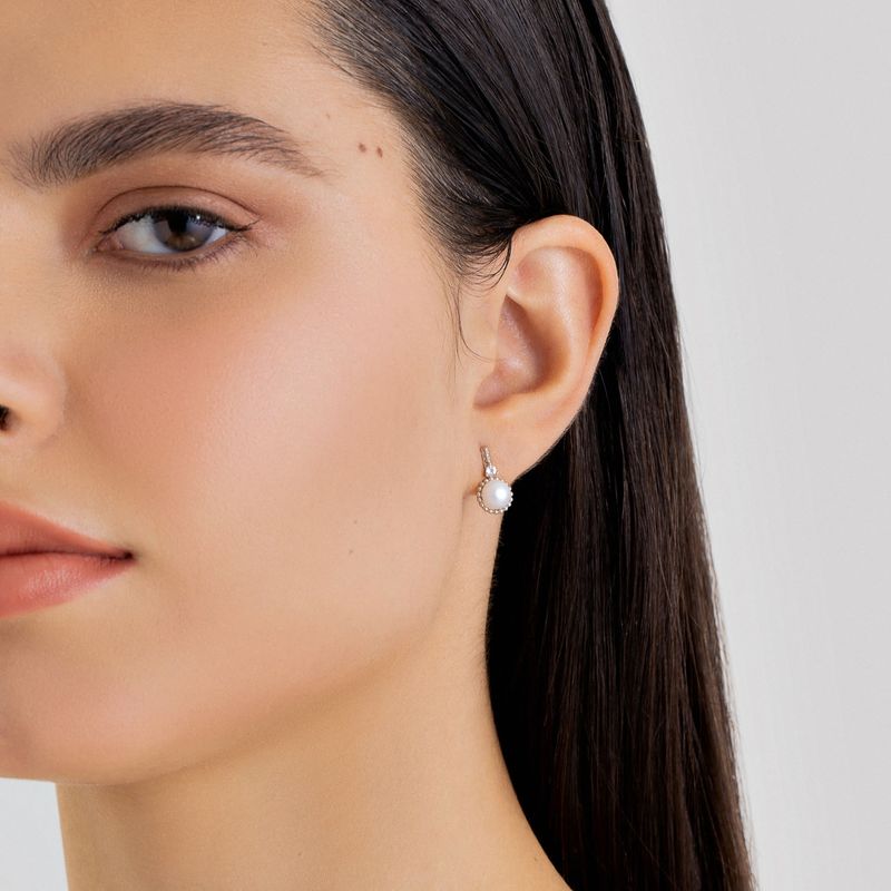 Brincos de prata com pérola e detalhes brilhantes, usados na orelha de uma mulher com cabelo castanho escuro.