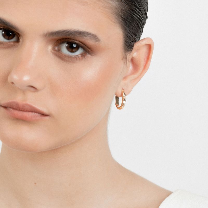 Mulher com brincos dourados de argola, usando maquiagem natural e cabelo escuro e liso, contra fundo branco.