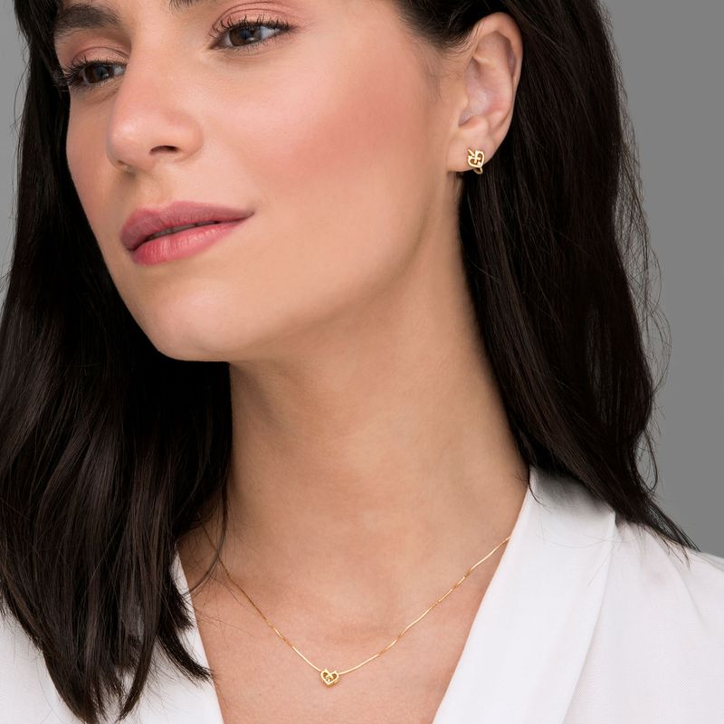 Close de uma mulher usando joias de ouro: um par de brincos e um colar delicado com um pequeno pingente em forma de coração.