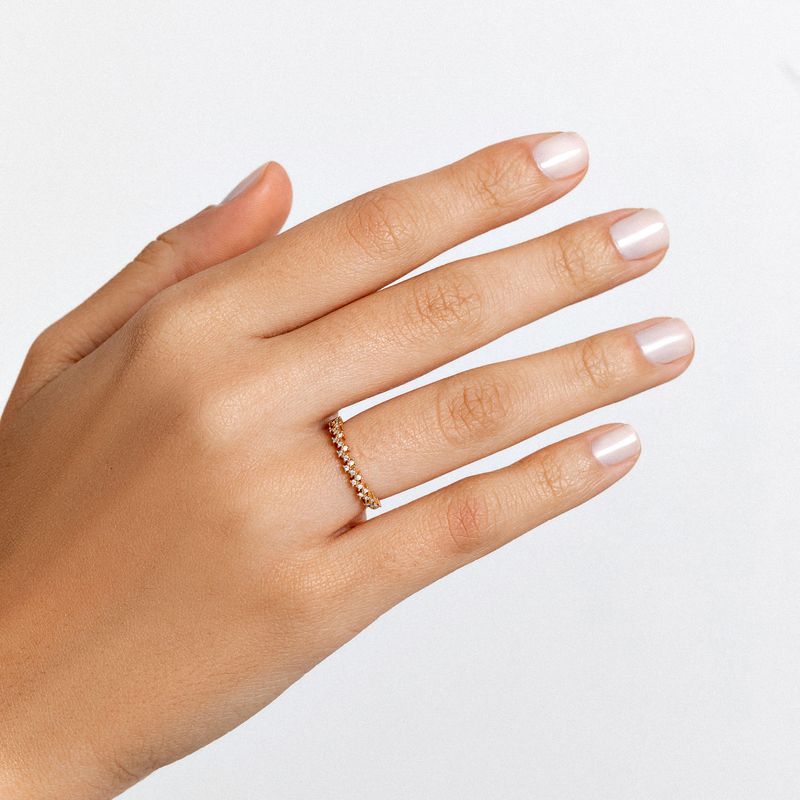 Mão com unha nude, usando um anel delicado com pedras brilhantes na posição do anelar. Fundo branco.