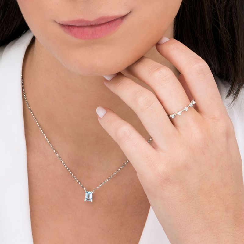 Mulher usando colar com pedra azul e anel prateado com pedras transparentes na mão direita, perto do rosto.