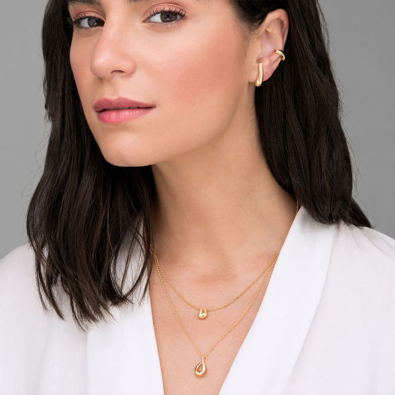Mulher usando três colares dourados finos de diferentes comprimentos e três brincos dourados na orelha direita, com cabelo castanho escuro e roupa branca.