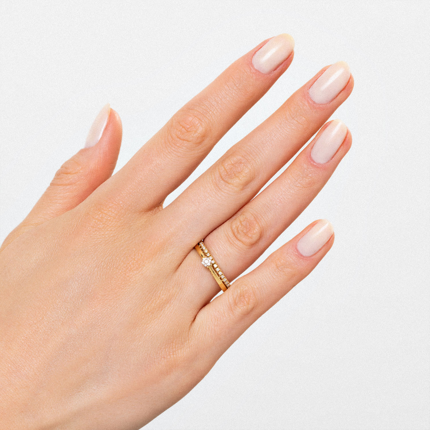 Mão com unhas nude e um anel dourado com pedras brilhantes na argola do dedo indicador.