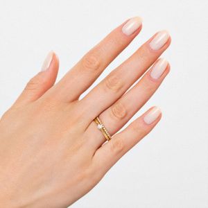 Mão com unhas nude e um anel dourado com pedras brilhantes na argola do dedo indicador.