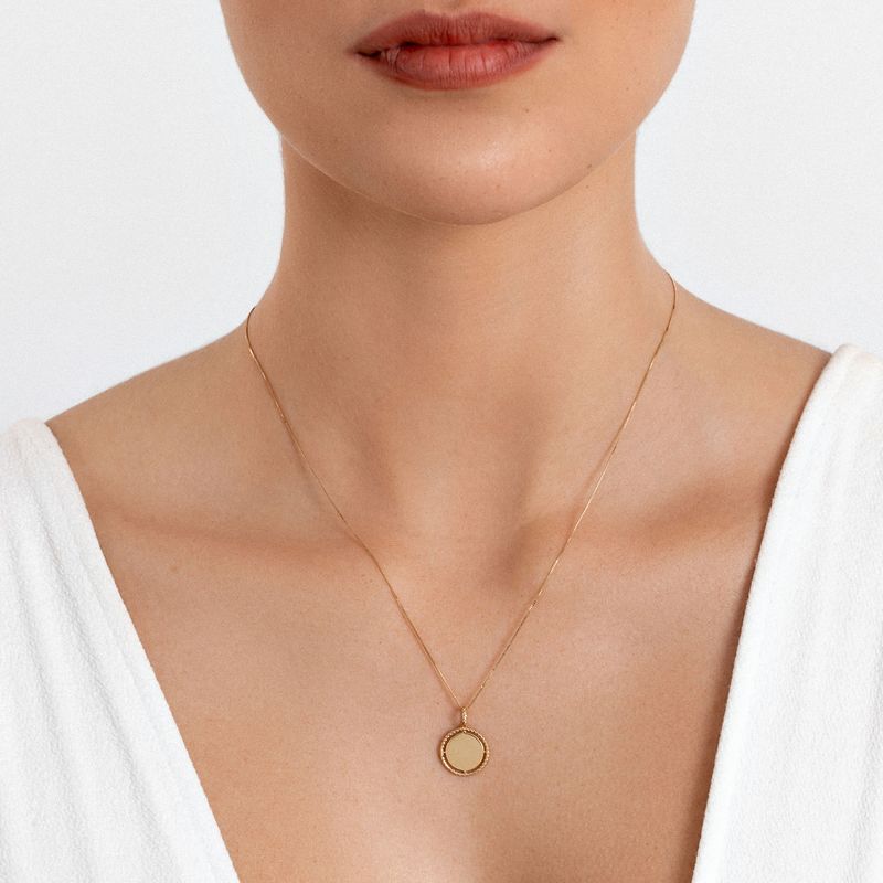 Colar dourado com pingente circular simples, usado por uma pessoa com blusa branca.