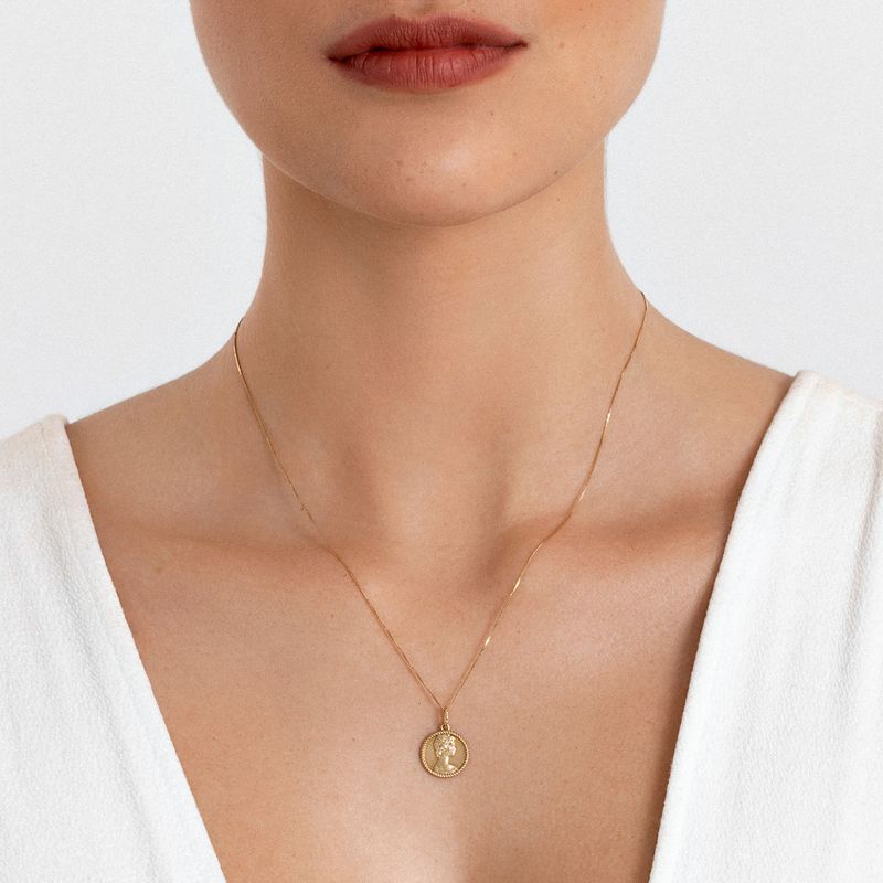 Colar dourado com pingente circular e árvore da vida, usado por uma pessoa com blusa branca.