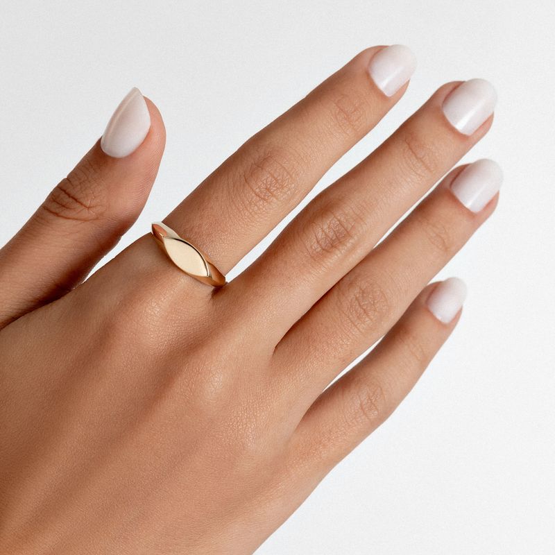 Mão com unhas brancas e um anel dourado em formato de folha na dedo médio, contra fundo branco.