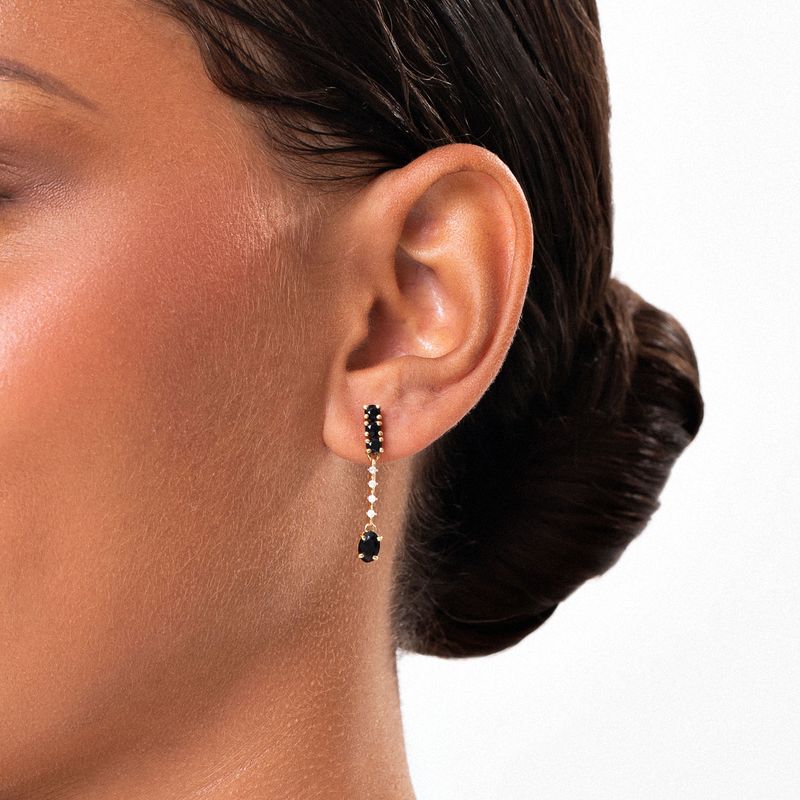 Brinco de argola longo com pedra preta e detalhes dourados na orelha de mulher com cabelo preso.