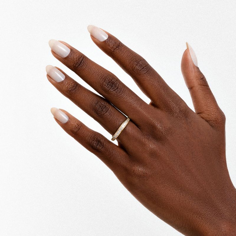 Mão com pele morena, unhas quadradas com esmalte nude, usando anel dourado com cristais na dedo médio.