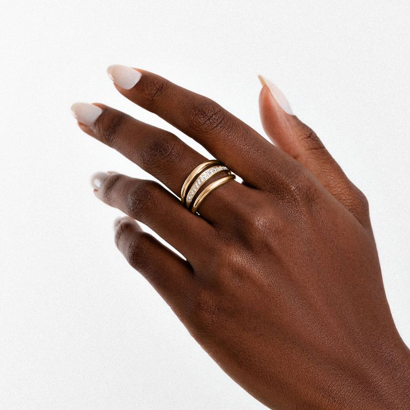 Mão com pele escura usando três anéis dourados com detalhes brilhantes, dedos alongados e unhas claras, contra fundo branco.