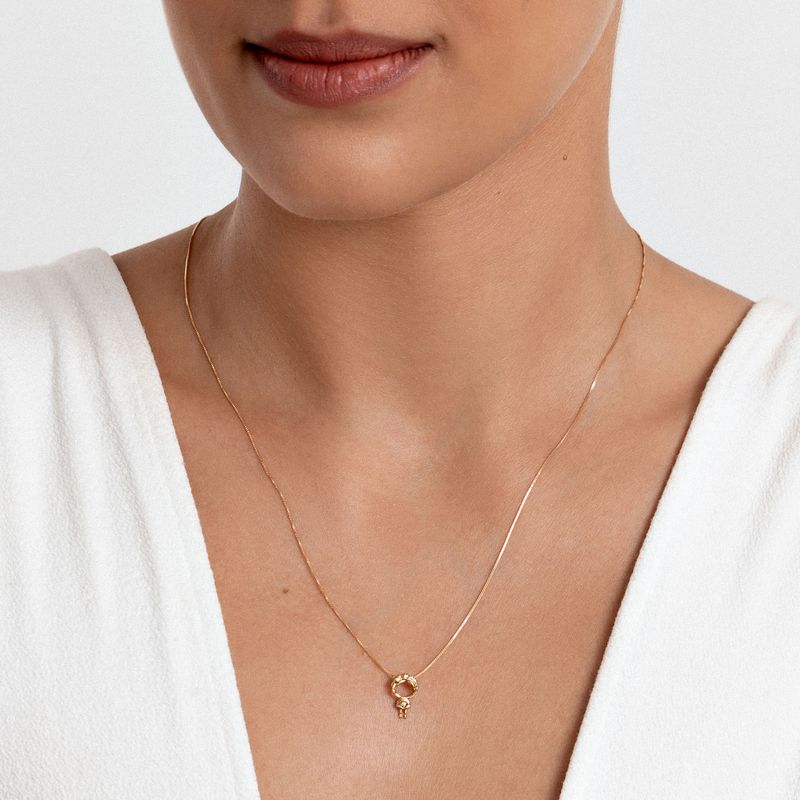 Colar dourado delicado com pingente circular com detalhes em zircônia, usado por uma mulher com blusa branca.